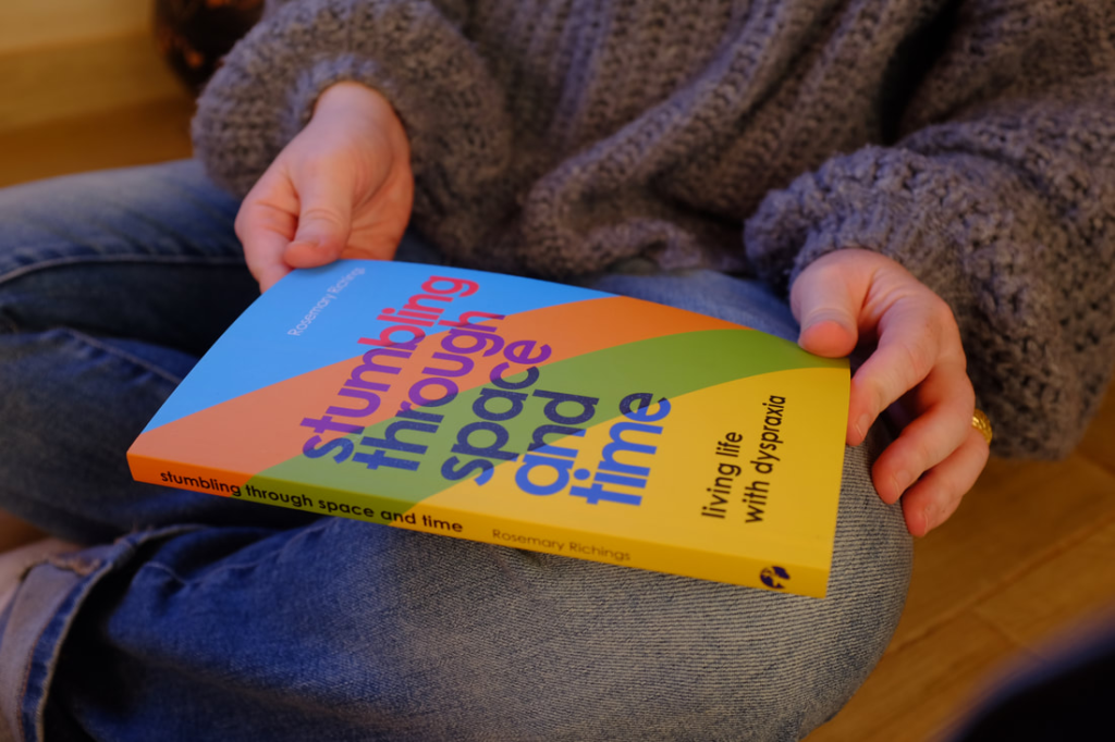 Cover of Rosemary Richings book Stumbling Through Space and Time: Living Life With Dyspraxia, held on her lap and on a side angle.