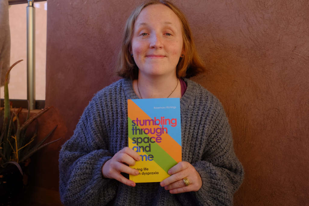 Rosemary Richings in a grey sweater holding up a copy of her book Stumbling Through Space and Time: Living Life With Dyspraxia. She is stood in front of a red wall. This photo has had its fair share of usages in press projects.