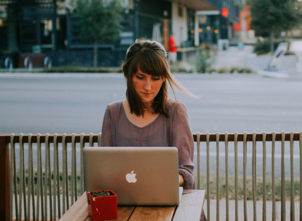 “Woman sitting at an outdoor table using a laptop, focused on the screen, with a street and buildings in the background.