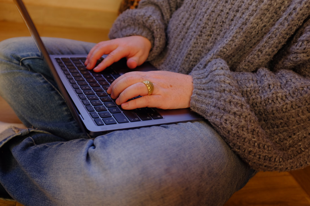 Close-up of Rosemary Richings sitting cross-legged while typing on a laptop, wearing a chunky grey knit sweater and blue jeans, with a gold ring on one finger. She is working on her latest neurodiversity themed project.