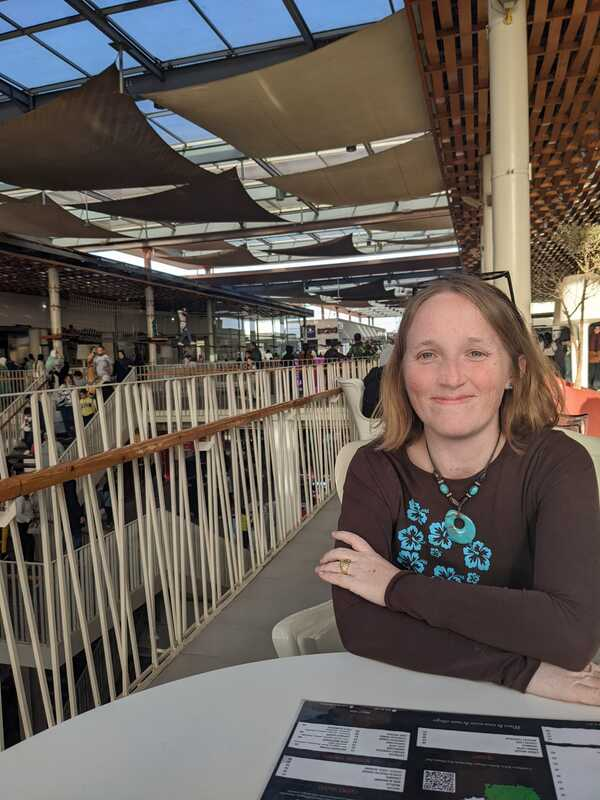 Photo of Rosemary Richings in brown shirt with blue flowers. She is sitting at a table smiling with her sunglasses on. This photo has been used a lot in press.