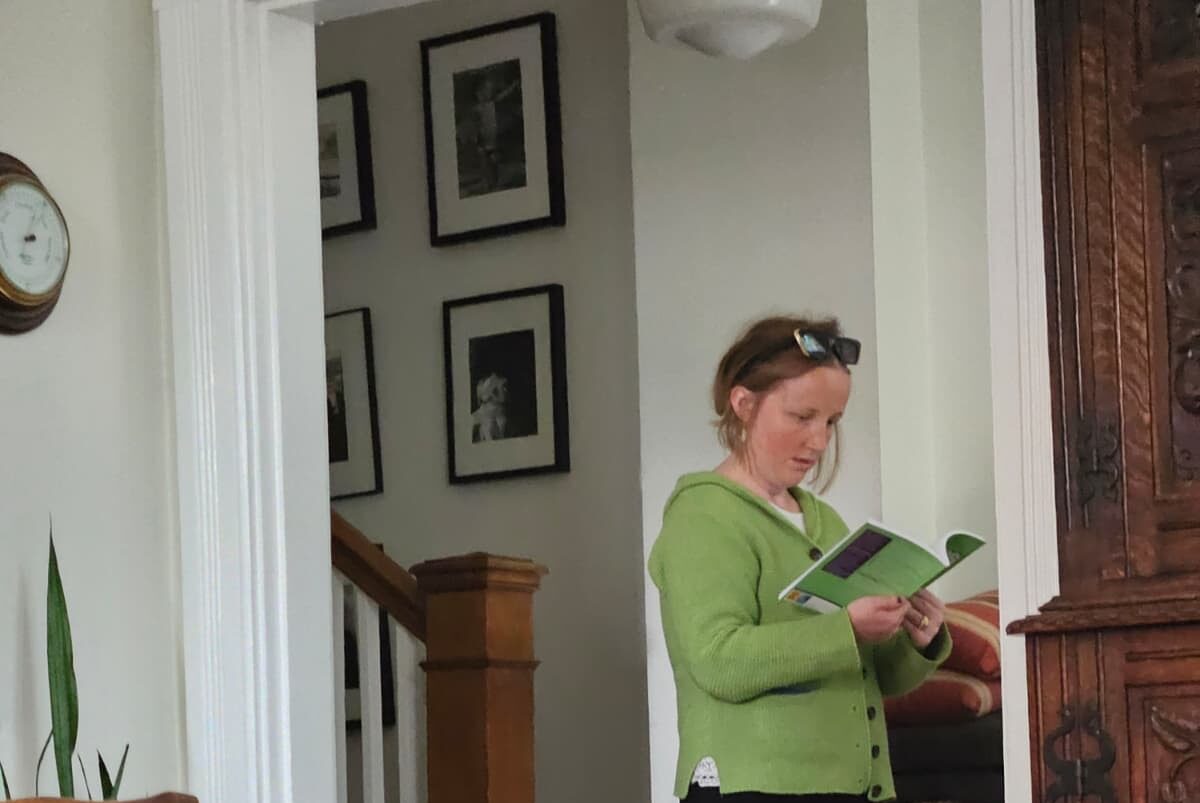 Author Rosemary Richings stands indoors reading her book “Studying With Dyspraxia.” She wears a green cardigan, black‑and‑white skirt, and tan sandals, with sunglasses resting on her head. The cozy home setting includes a staircase, framed black‑and‑white photographs, a wooden cabinet, a potted plant, a patterned rug, and a wall clock, capturing a warm everyday moment of engaging with her own work.