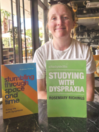 Author Rosemary Richings sits at a café table with her two books displayed in front of her: “Stumbling Through Space and Time: Living Life With Dyspraxia” and “Studying With Dyspraxia.” The background shows tables, chairs, hanging lights, and large windows filling the space with natural light, creating a welcoming setting for her work.